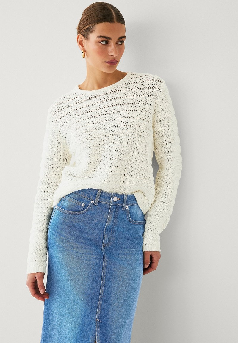 Maglione bianco lavorato a maglia con un motivo testurizzato e ampio scollo rotondo, abbinato a una gonna di jeans blu chiaro con uno spacco frontale e chiusura con bottoni.