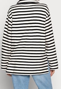 Schwarz-weiße gestreifte Fleecejacke mit einem Reverskragen, langen Ärmeln und seitlichen Schlitzen, aus einem weichen, strukturierten Stoff gefertigt.
