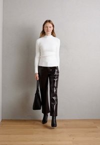 Weißer, langärmliger, taillierter Strickpullover kombiniert mit hochgeschnittenen, glänzenden schwarzen Lederhosen. Das Model trägt eine schwarze Tote-Tasche und schwarze Stiefeletten.