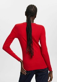 Red long-sleeve shirt with a crew neck, fitted design, and smooth texture, paired with blue jeans; hair styled in long braids.