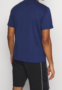 Muž oblečený v jednoduchém námořnickém modrém tričku s krátkým rukávem a černých sportovních šortkách s bílými bočními pruhy, postávající na neutrálním pozadí.