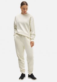 Sudadera color crema con un ajuste relajado y puños acanalados, combinada con joggers a juego que tienen una cintura elástica. Zapatillas negras.