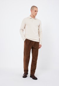 Polo beige a maniche lunghe con tre bottoni, abbinato a pantaloni in velluto a coste marrone. Modello che indossa stivali alla caviglia marrone scuro. Design semplice e casual.