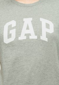 Tissu gris clair avec de grandes lettres majuscules blanches "GAP" imprimées en travers de la poitrine dans une police audacieuse et carrée.