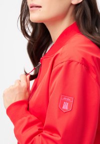 Röd bomberjacka med dragkedja fram, ribbade ärmslut och en texturerad patch med ett slott-logotyp på vänster ärm. Mjuk tygfinish.