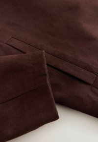 Tissu en daim marron foncé montrant un gros plan d'un poignet de manche et d'une poche fendue avec des coutures nettes.