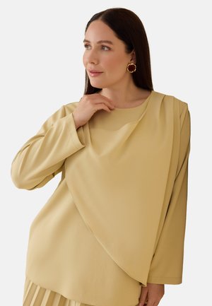 Kvinde med langt mørkt hår iført beige lagdelt top og guldkæder, rører ved sin krave, mens hun ser mod højre på en ensfarvet baggrund.