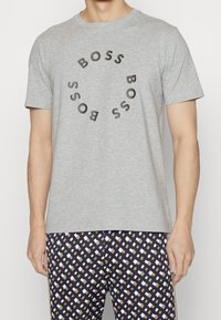 Muž v světle šedém tričku s krátkým rukávem, na kterém je kulatý nápis "BOSS", a v patterned černých, béžových a bílých kalhotách.