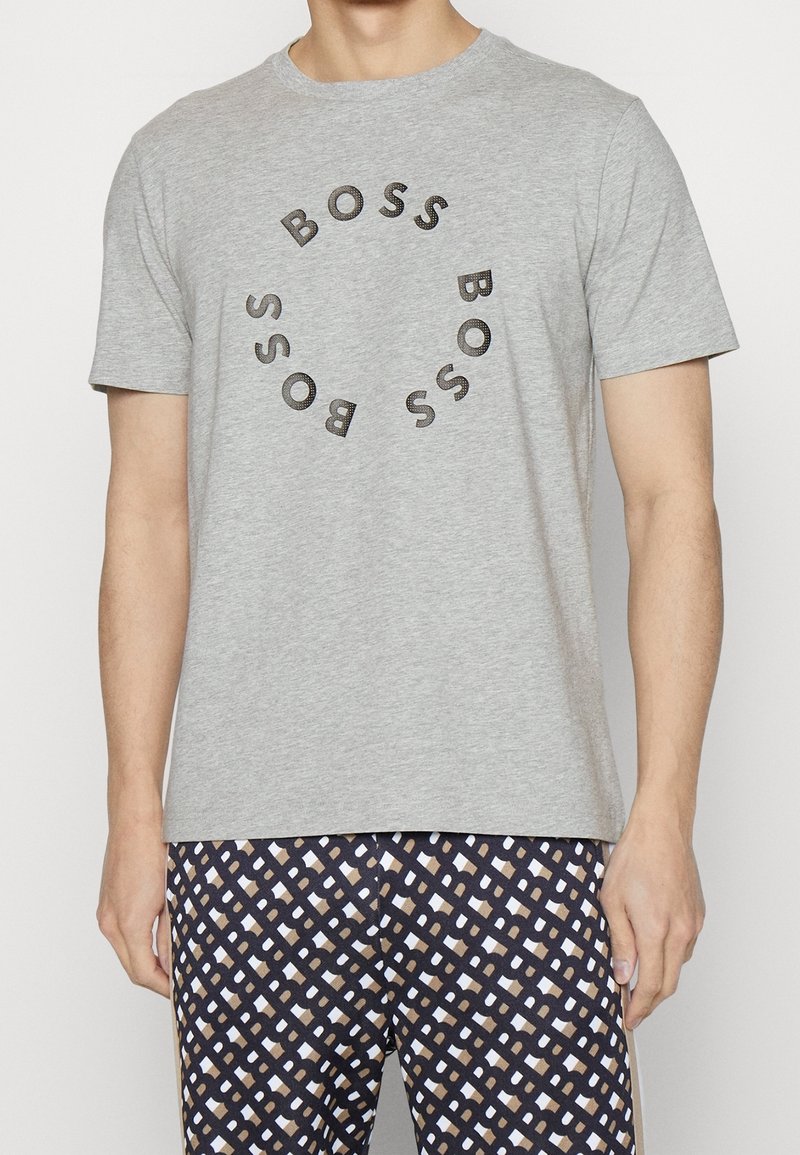 Muž v světle šedém tričku s krátkým rukávem, na kterém je kulatý nápis "BOSS", a v patterned černých, béžových a bílých kalhotách.