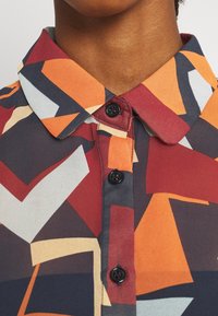 Camicia colorata e decorata con un design rosso, arancione e blu navy. Presenta un colletto e bottoni neri, realizzata in un tessuto morbido e leggero.