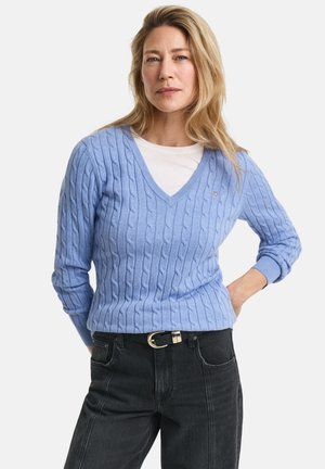 Lyseblå kabelstrikket genser med V-hals og lange ermer, kombinert med en hvit skjorte under og sorte høytlivsjeans.