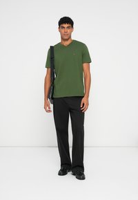 Πράσινο μπλουζάκι v-neck από βαμβάκι, σε συνδυασμό με μαύρο παντελόνι και παπούτσια. Το μοντέλο στέκεται μπροστά σε λευκό φόντο, κρατώντας μια τσάντα.