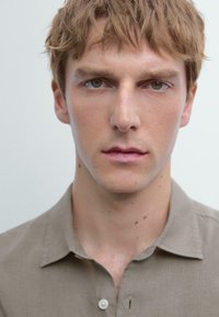 Close-up portrait of a person with short, light brown hair. Wearing a light beige button-up shirt with a collar and visible stitching.