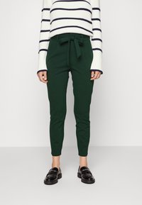 Zielone dopasowane spodnie z detalem w pasie na sznurku, wykonane z gładkiego materiału, w połączeniu z białym swetrem w paski. Czarne mokasyny dopełniają stylizację.