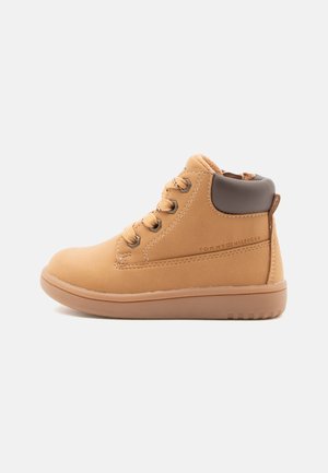 Bottes montantes en cuir synthétique beige avec des accents marron, bout rond, design à lacets et semelle en caoutchouc texturé. Logo de la marque sur le côté.