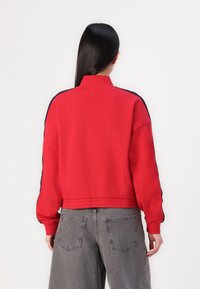 Felpa rossa corta con collo alto; presenta strisce blu lungo le spalle e i polsini; morbido al tatto; abbinata a jeans grigi a vestibilità ampia.