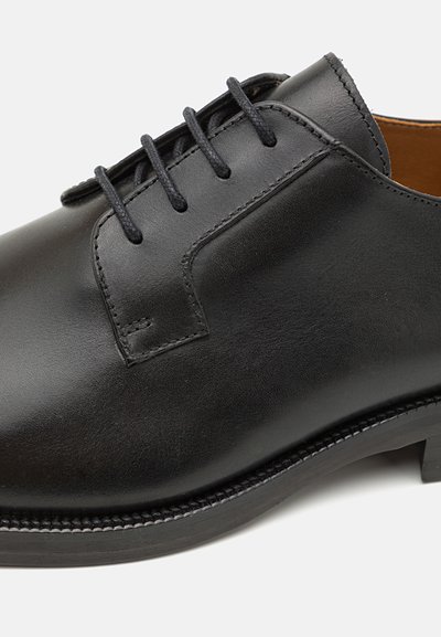 Scarpa nera in pelle con lacci, dalla texture liscia, punta arrotondata e dettagli di cucitura minimi. Include lacci neri e suola piatta in gomma.