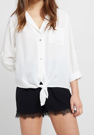Chemise blanche légère à boutons avec une poche, dotée d'un détail de nouage à l'avant. Associée à un short noir orné de bordure en dentelle.