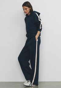 Woman wearing navy blue hoodie and matching wide-leg pants with beige side stripes, standing with hands in pockets against white wall.