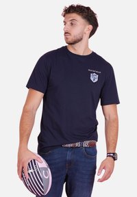 T-shirt en coton bleu marine avec un logo de club de rugby sur la poitrine gauche ; associé à un jean en denim et tenant un ballon de rugby rayé.