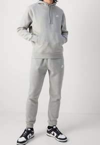 Felpa grigia con tasca frontale e coulisse, abbinata a pantaloni felpa grigi coordinati. Entrambi presentano un piccolo logo Nike bianco. Sneakers in bianco e nero completano il look.