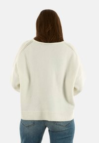 Persona con lunghi capelli castani che indossa un ampio maglione bianco a maglia e jeans blu, in piedi con la schiena rivolta verso un sfondo neutro.