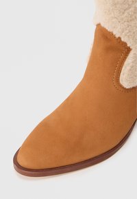 Bottino alla caviglia in suede beige con punta appuntita. La parte superiore presenta un morbido bordo in shearling di colore crema e una superficie strutturata.