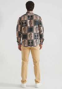 Chemise à manches longues avec un motif géométrique noir et beige, présentant des formes et des designs variés ; assortie à un pantalon beige et des baskets blanches.