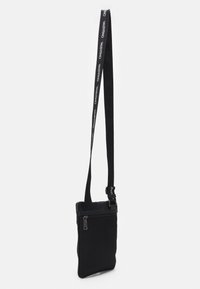 Sac bandoulière en nylon noir avec un compartiment principal zippé, une sangle ajustable et un logo discret sur la sangle. Texture lisse, design slim.