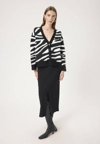 Cardigan zebra cu dungi negre și albe, realizat dintr-un material texturat, cu decolteu în V, închidere cu nasturi și purtat peste o fustă ajustată, neagră.