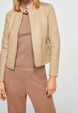 Vrouw in een beige leren jas over een mauve gebreide trui en bijpassende gebreide broek, staand met ontspannen armen naast zich.