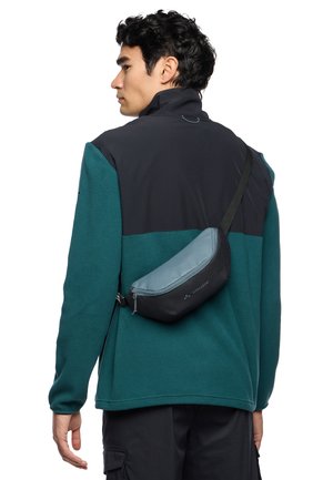 Schwarze und türkise Fleecejacke mit hohem Kragen, getragen über dunklen Hosen, ausgestattet mit einer kompakten, geschwungenen, grauen Umhängetasche mit Reißverschlussdetails.