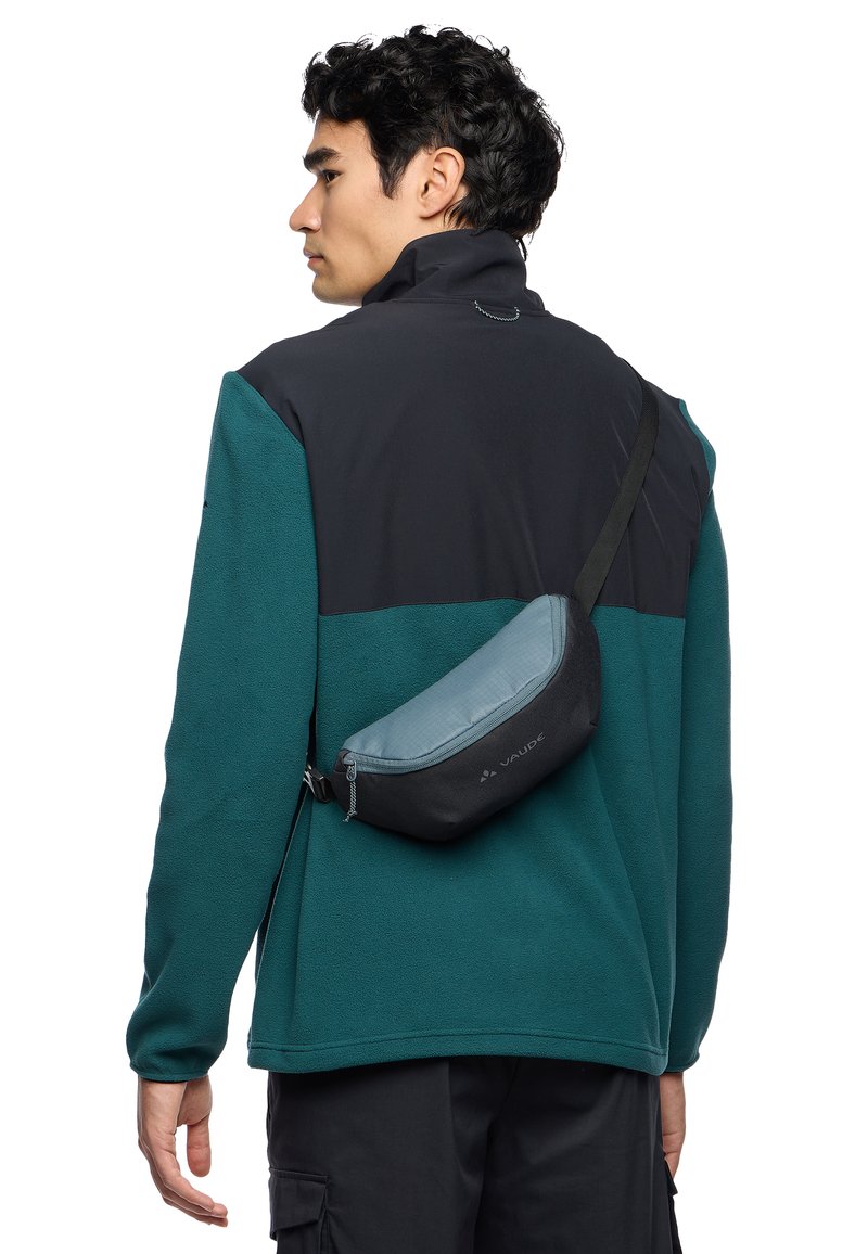 Veste en polaire noire et teal avec un col haut, portée sur un pantalon sombre, comporte un sac banane grisé compact et incurvé avec détail de fermeture éclair.