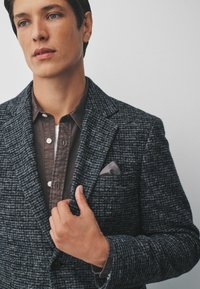 Sacou închis, țesut, cu un model texturat gri. Sub acesta, o cămașă maronie cu detalii cu nasturi. Borcea adaugă un accent elegant.