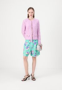 Pinker Spitzenjacke mit goldenen Knöpfen, kombiniert mit minzgrünen floralen Shorts und schwarzen Riemchen-Absätzen, und einer silbernen Handtasche haltend.