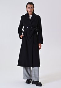 Schwarzer Wollmantel mit Schalkragen, tailliertem Gürtel und zwei Kn�öpfen. Das Modell trägt karierte Hosen und klobige schwarze Schuhe. Glatte Textur.