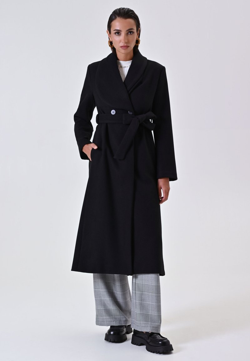Schwarzer Wollmantel mit Schalkragen, tailliertem Gürtel und zwei Knöpfen. Das Modell trägt karierte Hosen und klobige schwarze Schuhe. Glatte Textur.