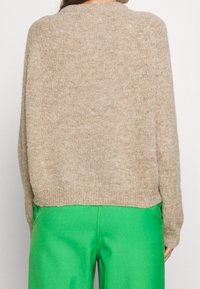 Persoon in een beige gebreide trui en felgroene broek, van achteren weergegeven tegen een effen witte achtergrond.