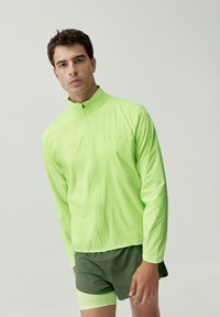 Giacca leggera, con zip completa, di un vivace verde lime, con colletto alto. Abbinata a pantaloni corti verdi scuri. Design semplice, senza motivi visibili o componenti metallici.