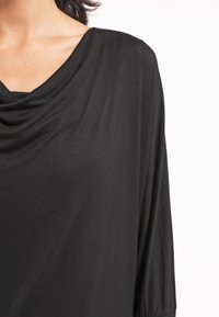 Femme portant un haut noir à encolure drapée, dévoilant l'épaule et le haut de la poitrine sur un fond blanc uni.