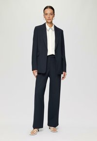 Marineblauer maßgeschneiderter Anzug mit einem strukturierten Blazer und weiten Hosen. Das Outfit umfasst eine weiße Bluse mit Kragen und einem dekorativen Kragen sowie Sandalen.