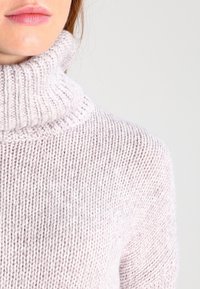 Ljusrosa stickad tröja med en hög, ribbad polokrage. Har en mjuk textur och lös passform som framhäver stickmönstret.