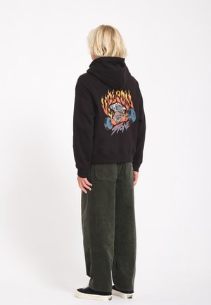 Zwarte hoodie met grafisch ontwerp van vlammen en een schedel, gecombineerd met groene corduroy broek. De capuchon is opgetrokken en de schoenen zijn zwarte sneakers.