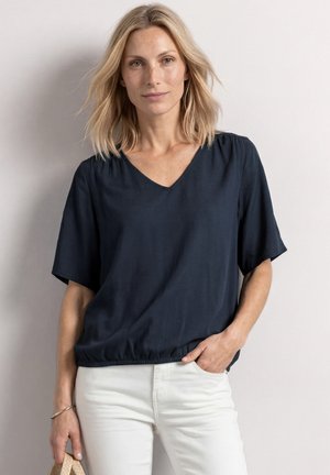 Femme blonde portant un haut ample bleu foncé à manches courtes et un pantalon blanc, debout avec une main dans la poche sur un fond uni.