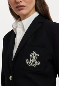 Blazer nero con una trama testurizzata, dotato di un logo ricamato bianco sulla tasca del petto e di un dettaglio con bottone argento.