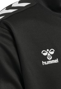 Zwarte sporttop van gladde stof, met een wit logo en een chevronpatroon op de schouders. Met een kortere halslijn.