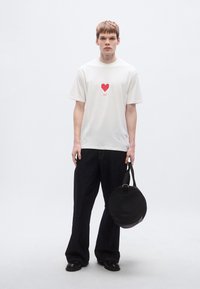 Λευκό κοντομάνικο μπλουζάκι με κόκκινη καρδιά και κείμενο "HUGO", συνδυασμένο με σκούρο τζιν φαρδιάς γραμμής και στρογγυλή μαύρη τσάντα. Ομαλή υφή, καθημερινό στυλ.