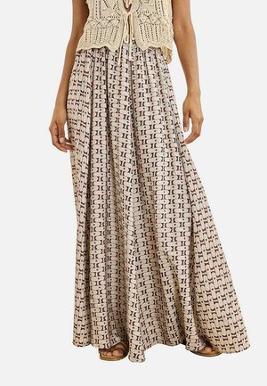 Mujer con una falda larga beige con estampado y un top sin mangas de crochet color crema, de pie con las manos relajadas a los lados.