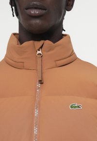 Brūna zip-up jaka ar augstu apkakli, kurai ir teksturēta auduma virsma un neliels izšūts krokodila logo uz krūšu daļas. Krāsains rāvējslēdzis pretējā krāsā.