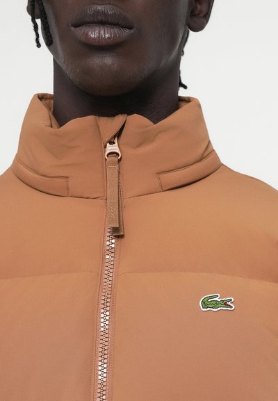 Veste zippée marron avec col haut, en tissu texturé et présentant un petit logo de crocodile brodé sur la poitrine. Fermeture éclair de couleur contrastante.